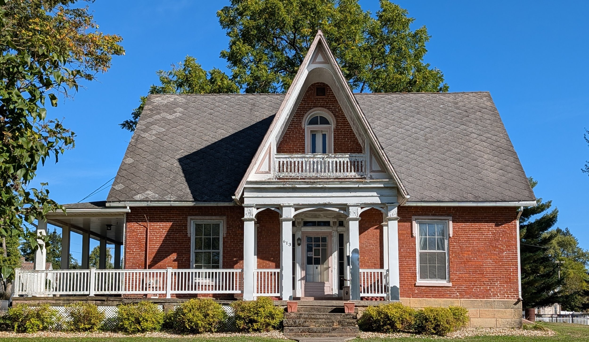 A two-story brick house with a steep gabled roof and white trim. It has a small porch with white railings and steps leading to the front door, and a balcony above the porch. The house has several windows and is surrounded by trees and a well-maintained garden.