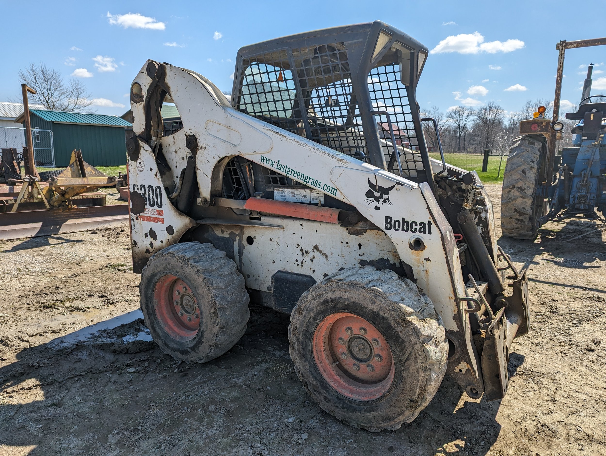 A skid-steer loader with a white body and black graphics, parked on a dirt surface under a blue sky with scattered clouds. There are trees and a green building in the background.