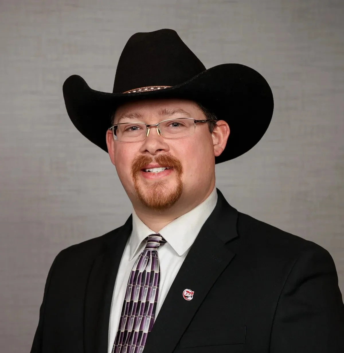 A man wearing glasses, a black cowboy hat, a suit, a patterned tie, and a lapel pin, smiling against a gray background.