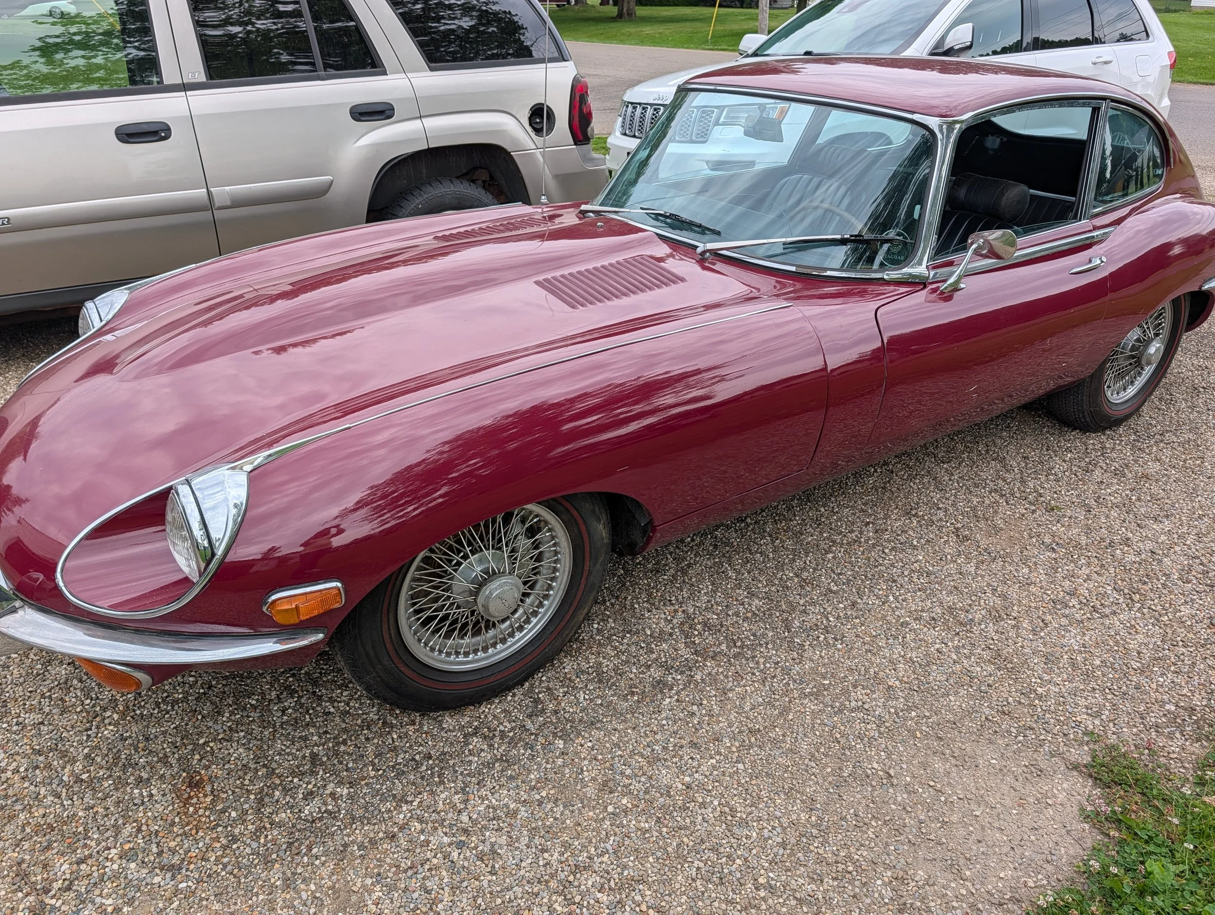 A vintage red Jaguar E-type with wire wheels parked on a gravel surface, with modern vehicles and green grass in the background.