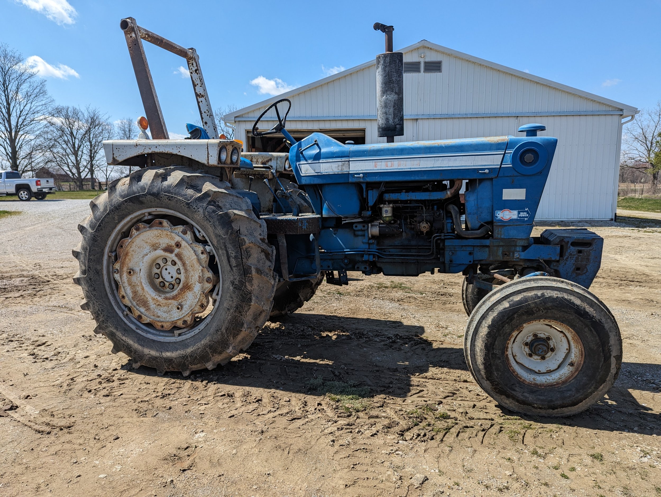 An old blue tractor with rust and dirt parked on dirt ground in front of a white building with a sloped roof. A pickup truck is visible in the background, and leafless trees are on either side of the building.