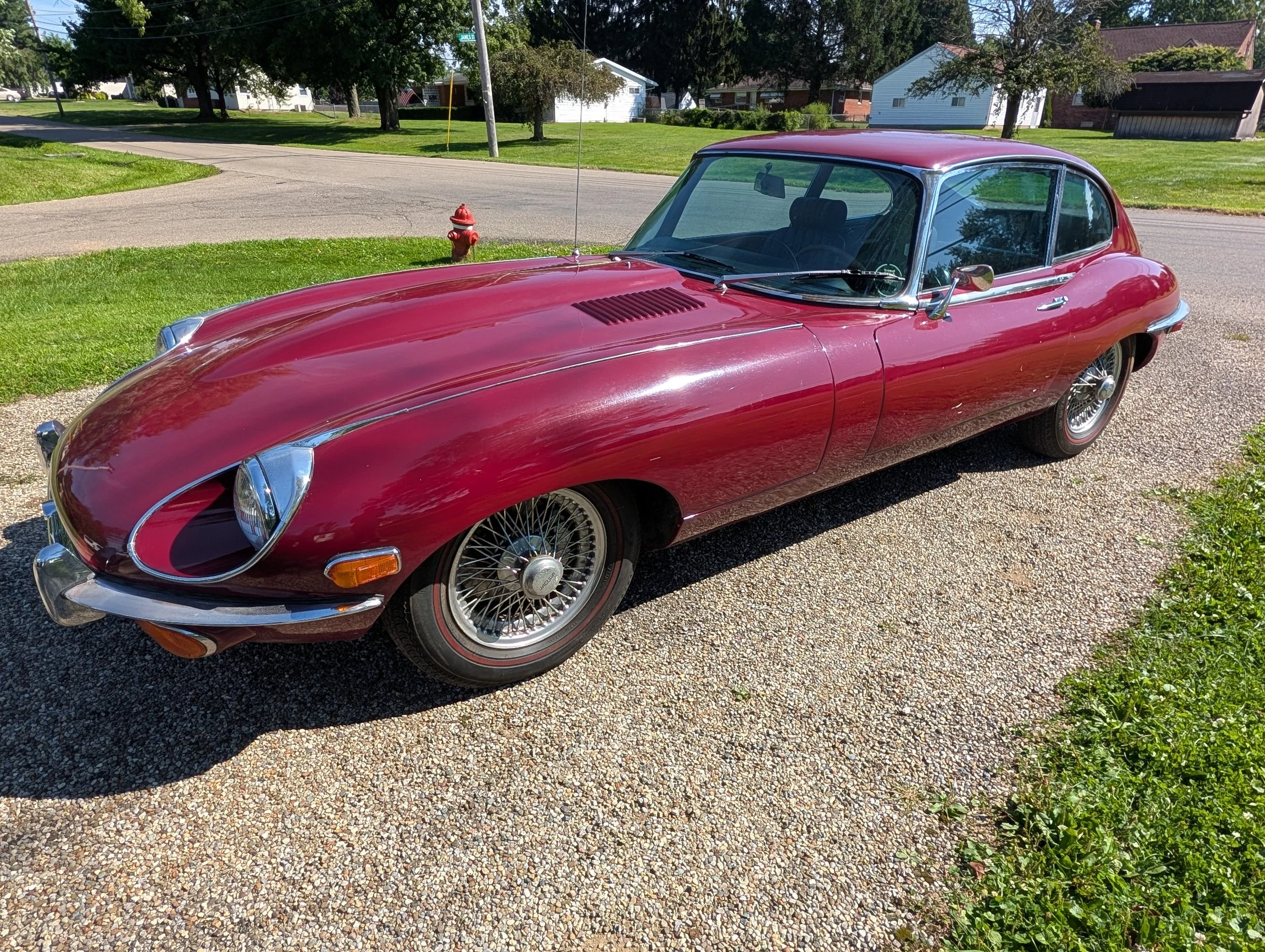 A vintage red Jaguar E-type with wire wheels parked on a gravel surface, with modern vehicles and green grass in the background.