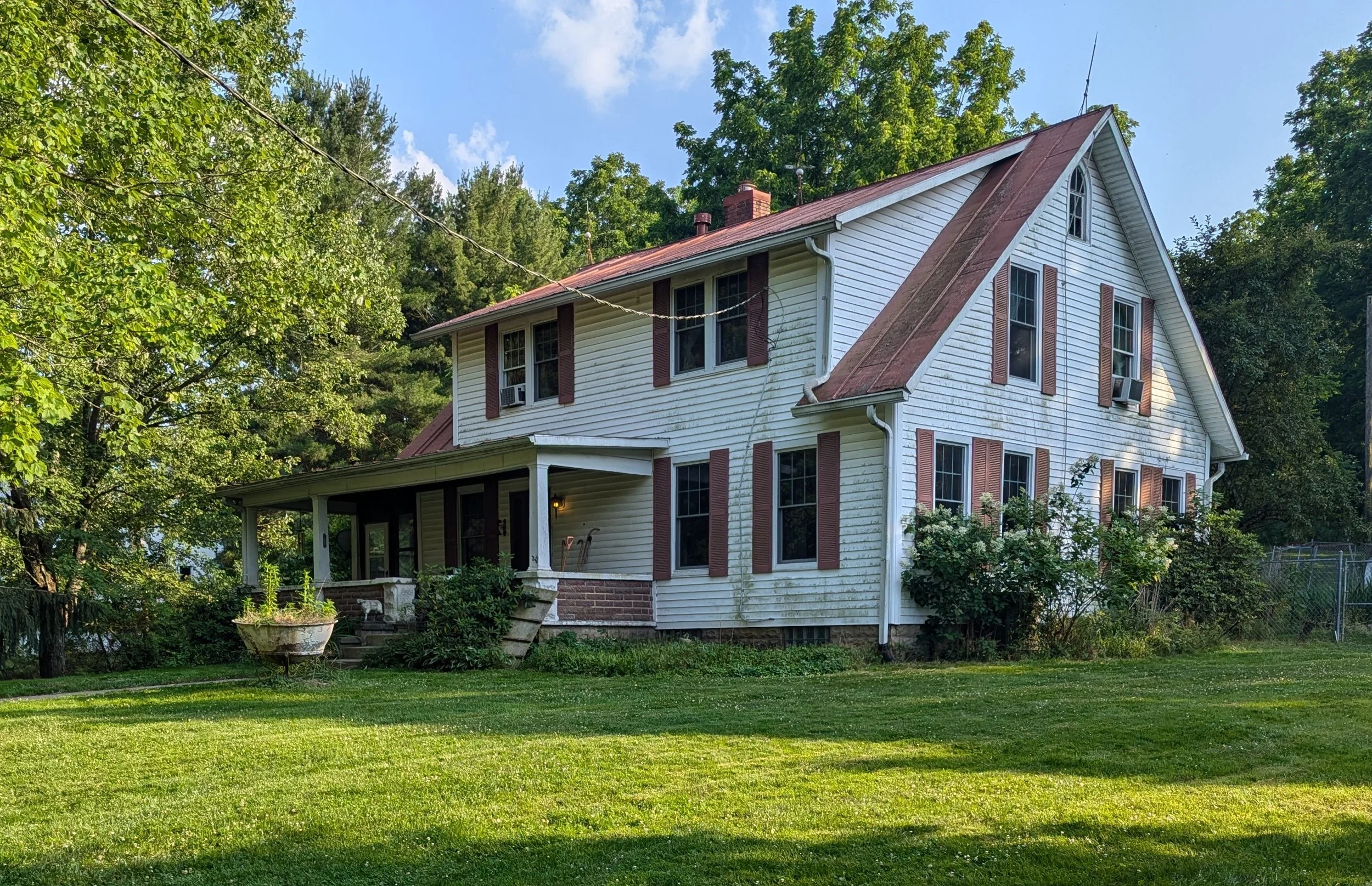 A two-story white house with brown shutters, a red roof, and a front porch is surrounded by green trees and a well-maintained grassy yard.