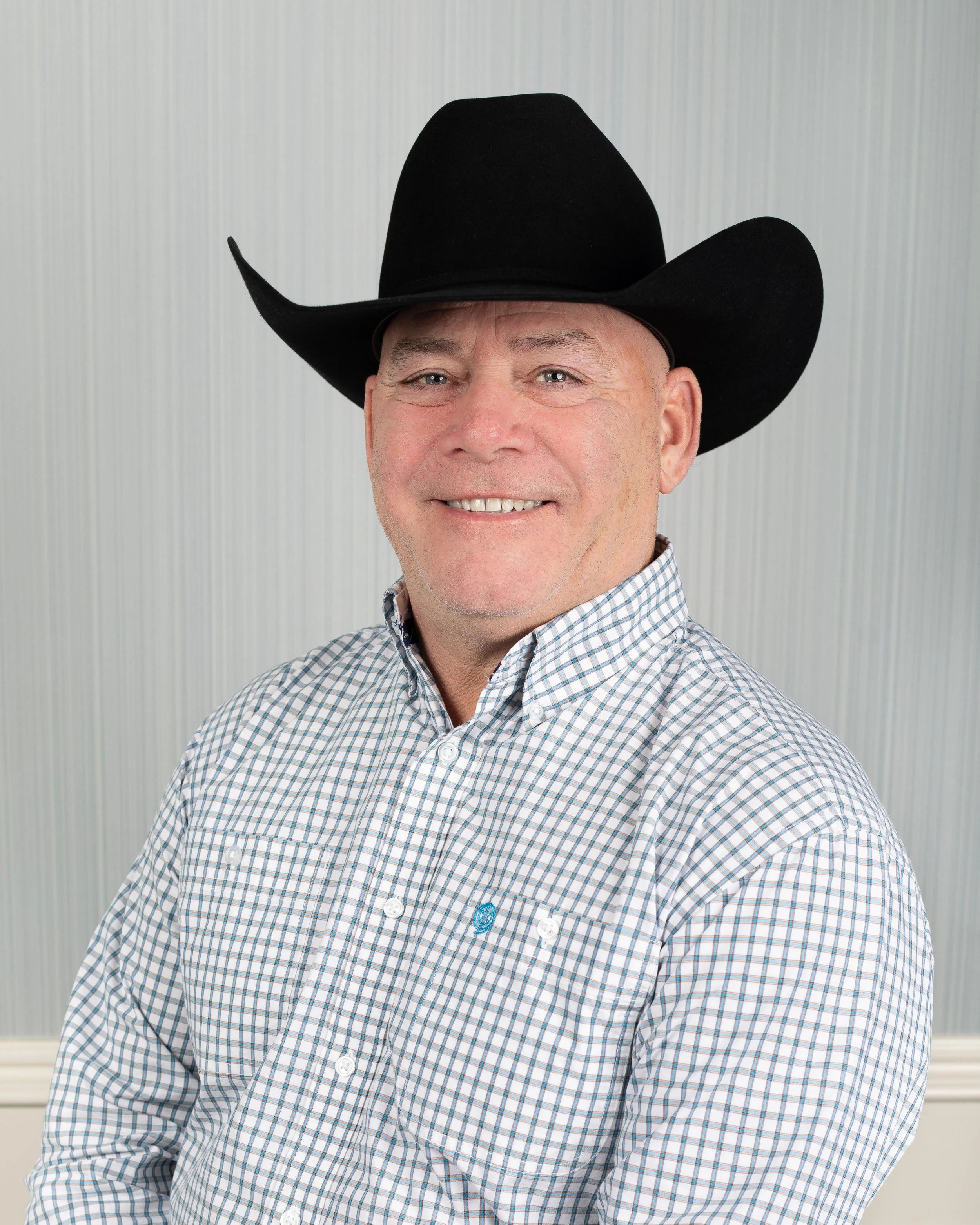 A smiling man wearing a black cowboy hat and a checked shirt standing in front of a plain background.
