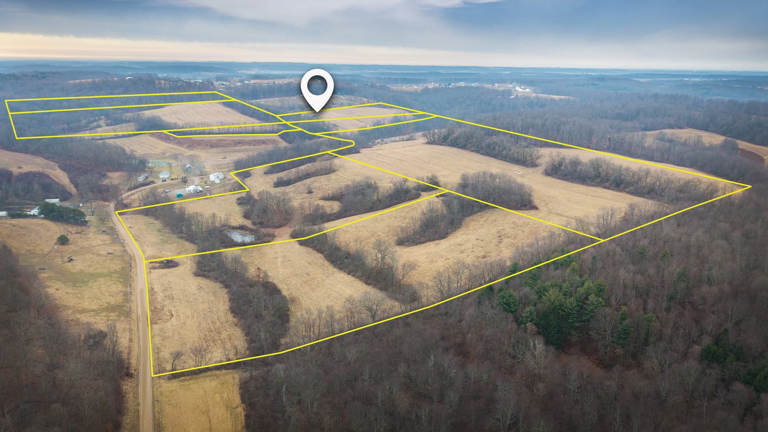 Aerial view of farmland with yellow boundary lines and a location pin marker overlay, showing fields, trees, and some buildings in a rural landscape.