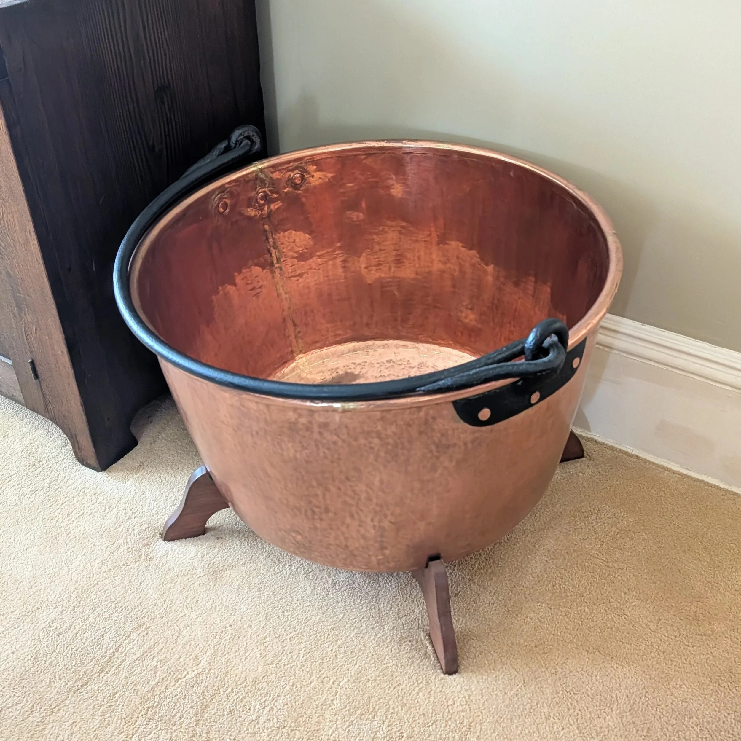 A wooden bowl with a dark finish and a rounded lip, sitting on a beige carpeted floor, with a piece of dark furniture in the background.
