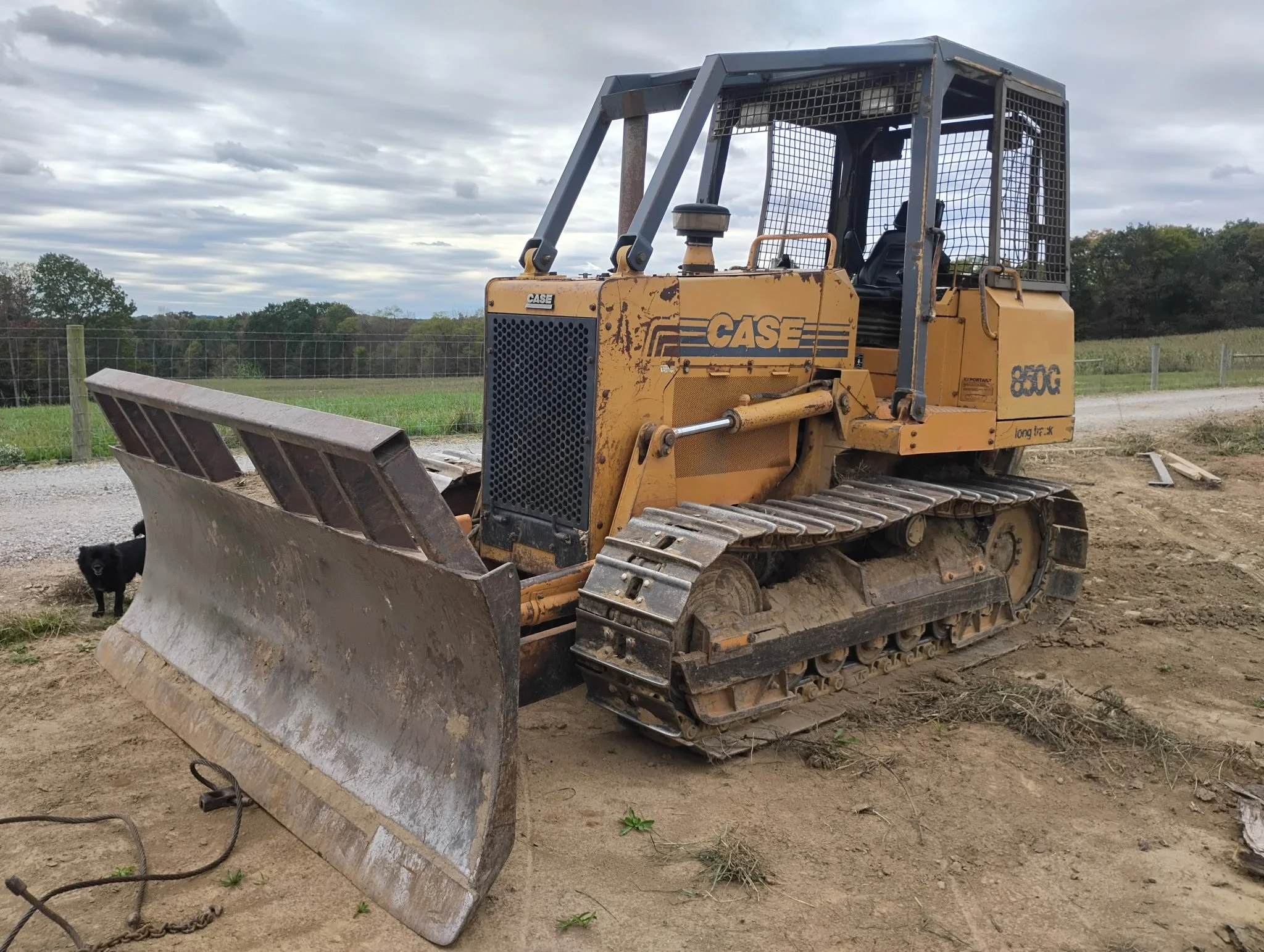 A yellow CASE bulldozer with a metal cage over the operator's seat parked on dirt ground in an open field area, with a cloudy sky overhead and trees in the background.