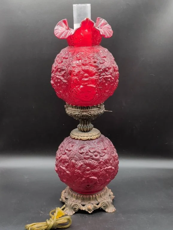 A vintage oil lamp with a glass chimney, featuring two ornate red glass globes with floral textures connected by a brass or bronze decorative element, resting on a carved metal base.