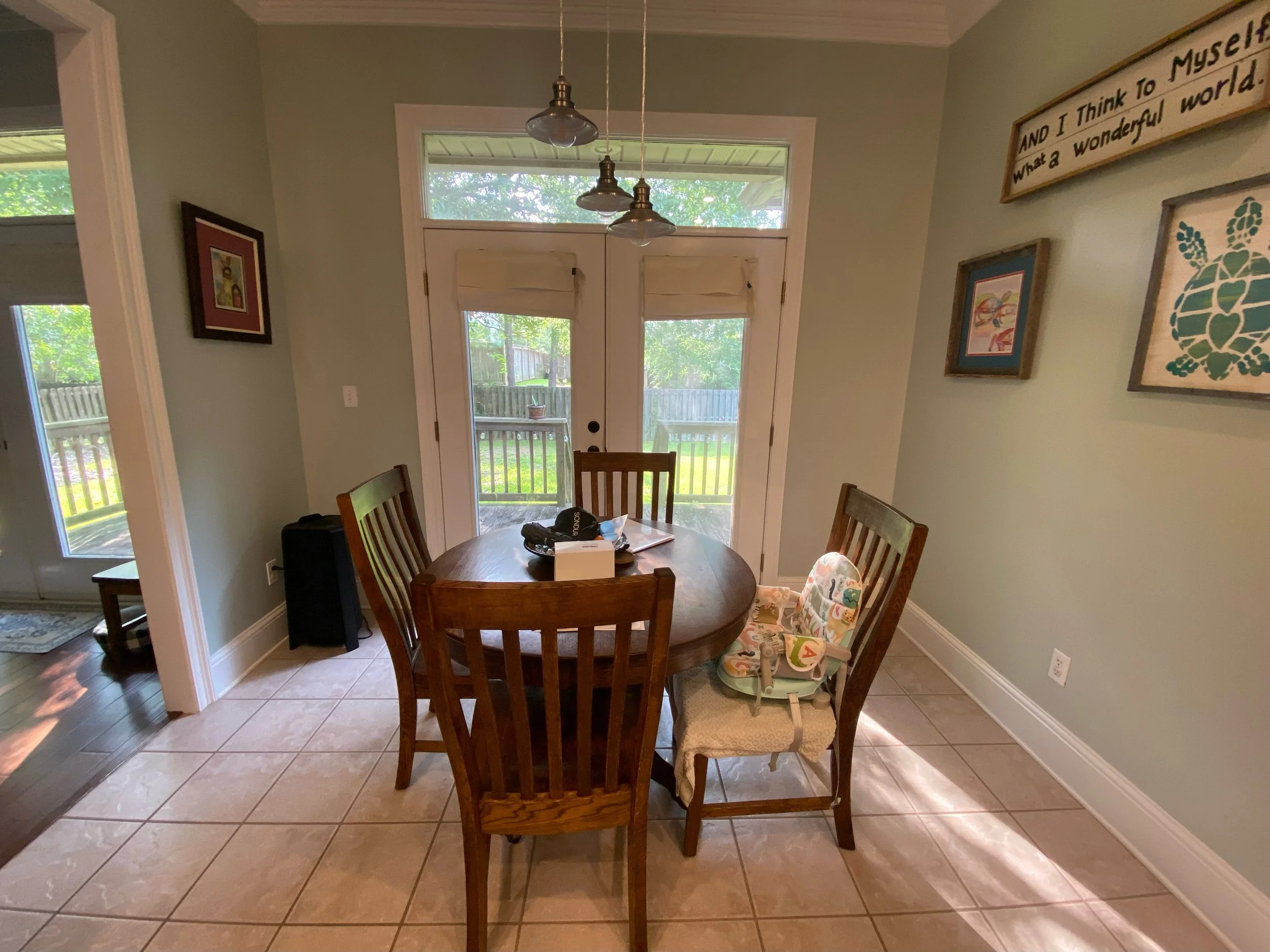 Dining area with a round wooden table, four chairs, and a high chair, with sliding glass doors leading to a backyard with a wooden fence and trees; hanging light fixtures above the table and artwork on the walls.