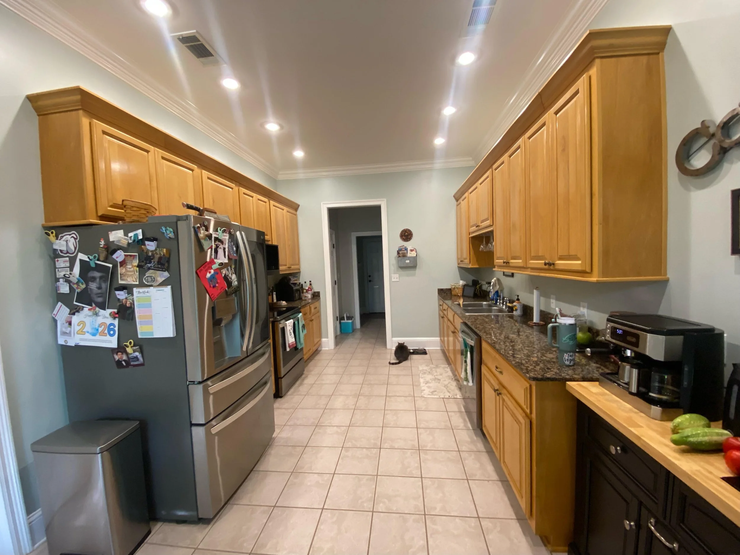 A kitchen with wooden cabinets, granite countertops, stainless steel appliances, a small black cat sitting on the tiled floor near the doorway, and various small items and appliances on the counters.