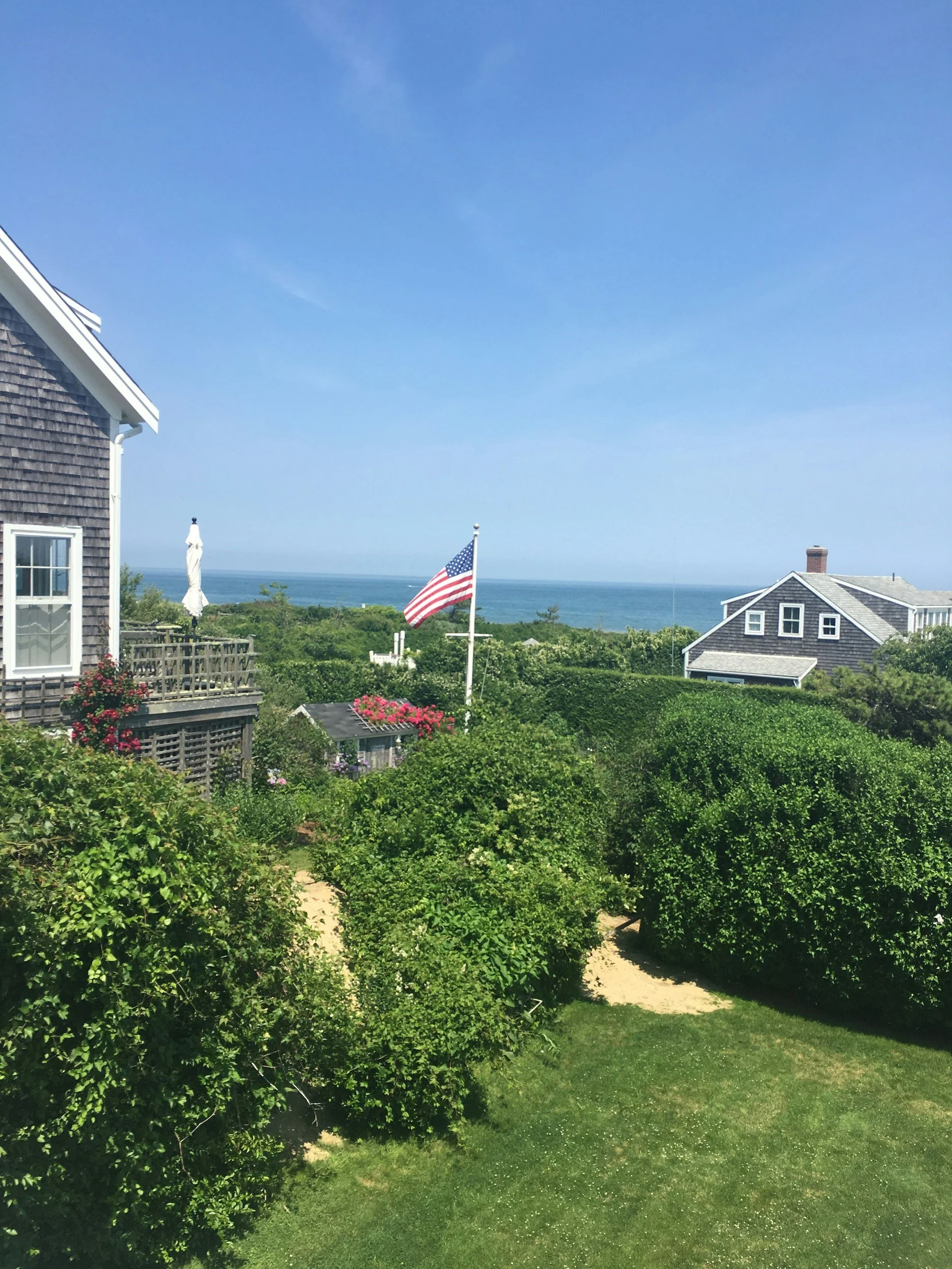 A backyard with greenery, including a garden and bushes. A flagpole with an American flag, houses with shingle siding, a patio umbrella, and the ocean in the background under a bright blue sky.