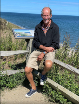 A man sitting on a wooden fence at a coastal overlook with ocean and blue sky in the background.