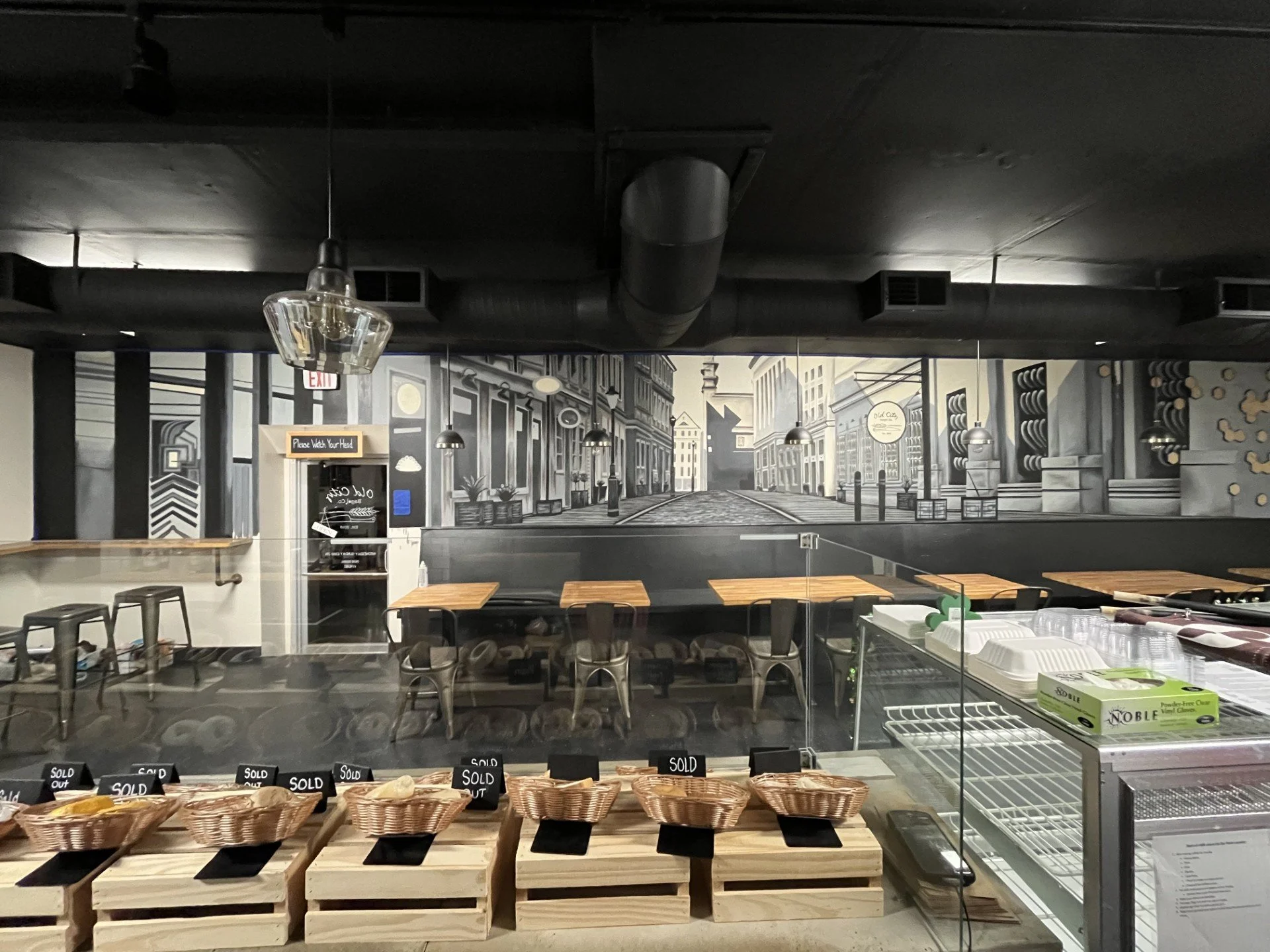 Interior view of a bakery or deli with a mural of a city street scene on the wall, wooden tables and chairs, baskets with baked goods marked as sold out, and a glass display case.