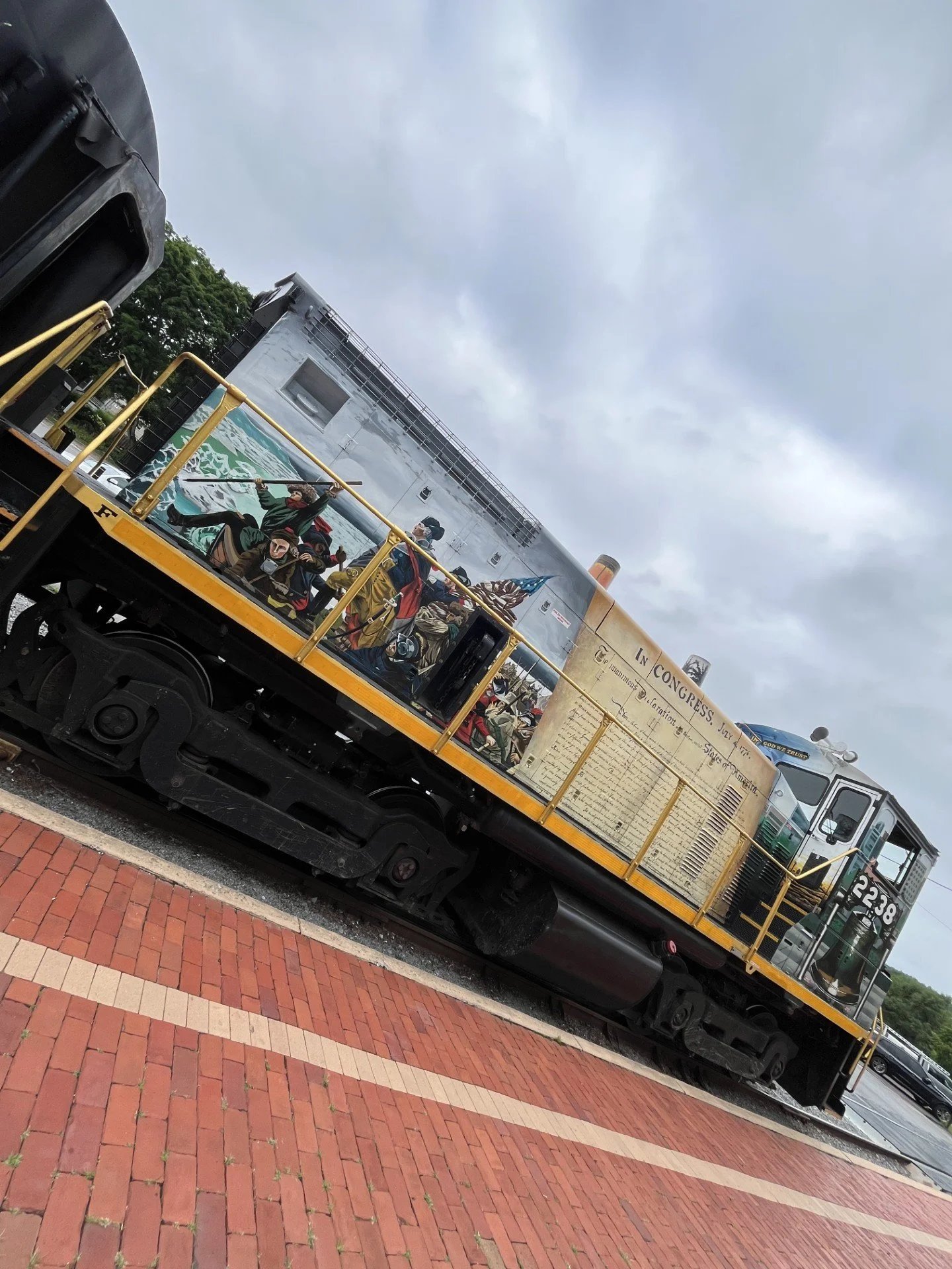 A train car decorated with historical images and text, parked on a brick-paved area with tracks, cloudy sky above, and some trees in the background.