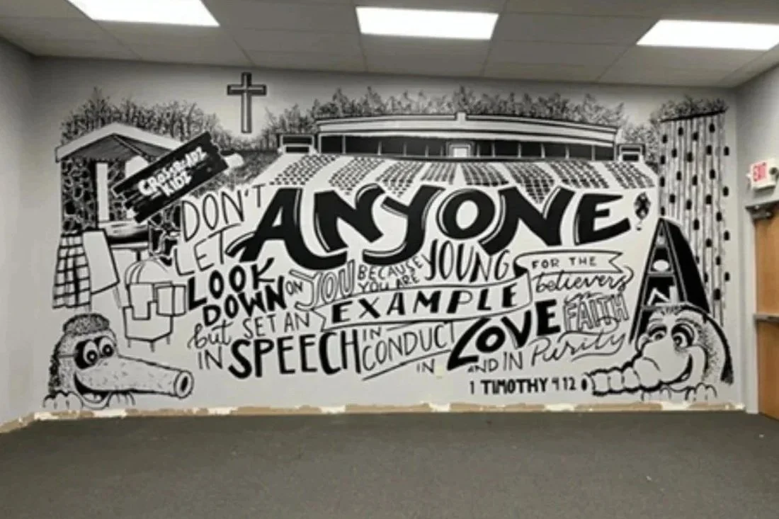 A church wall mural featuring a large biblical quote from 1 Timothy 4:12 in black and white, with cartoonish crocodile characters and a cross, decorated with trees and curtains in the background.