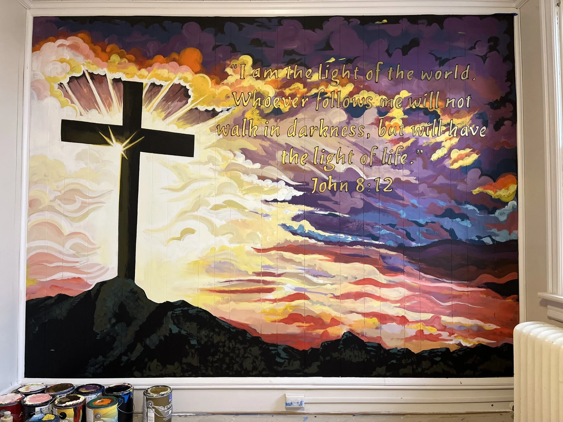 A large painted mural featuring a cross on a hill with a sunset and a vibrant, colorful sky filled with clouds and birds. The mural includes a biblical quote from John 8:12 about being the light of the world.