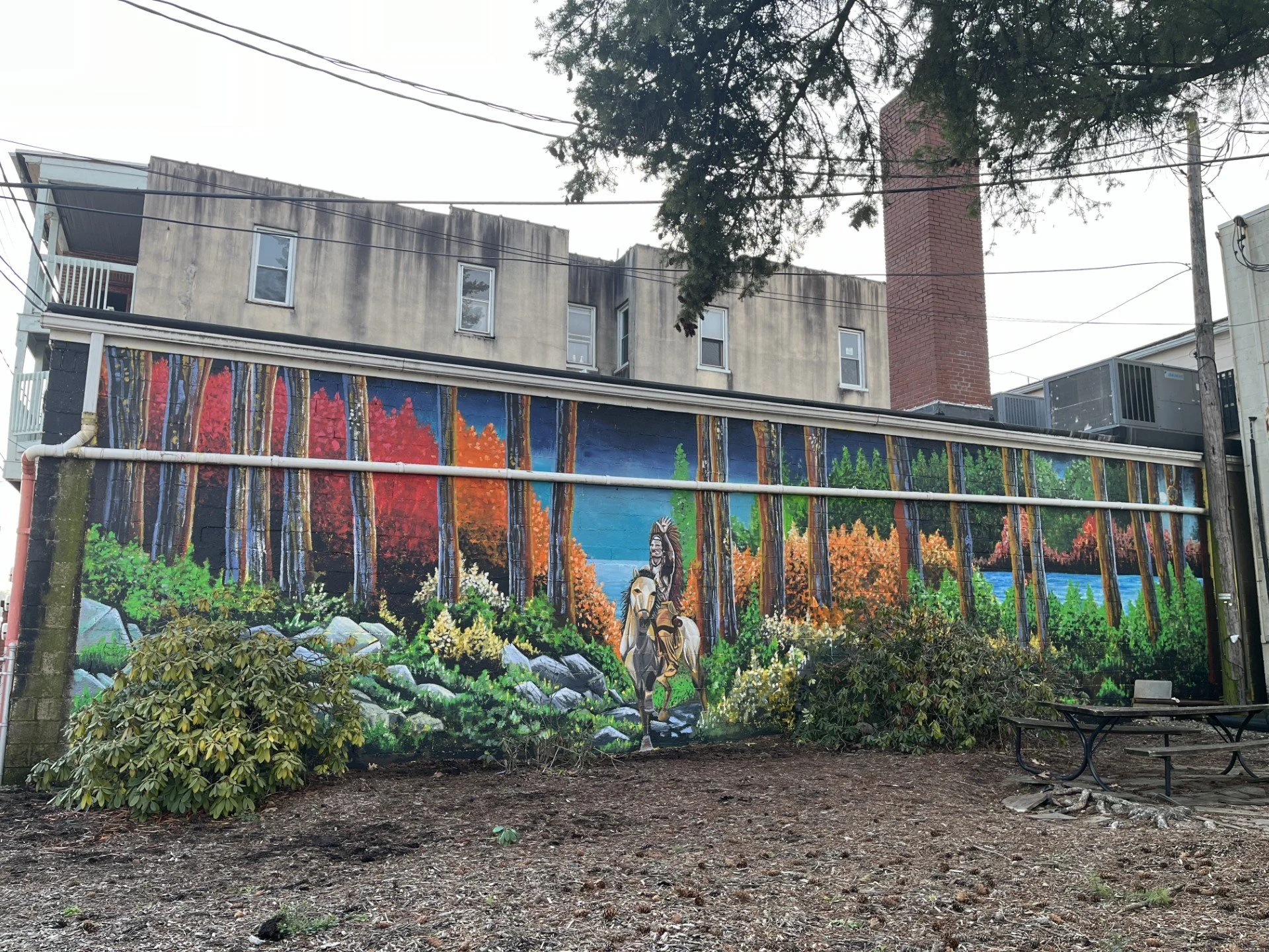 Colorful mural of trees, mountains, and a Native American figure in a park with benches and bushes, outdoor setting