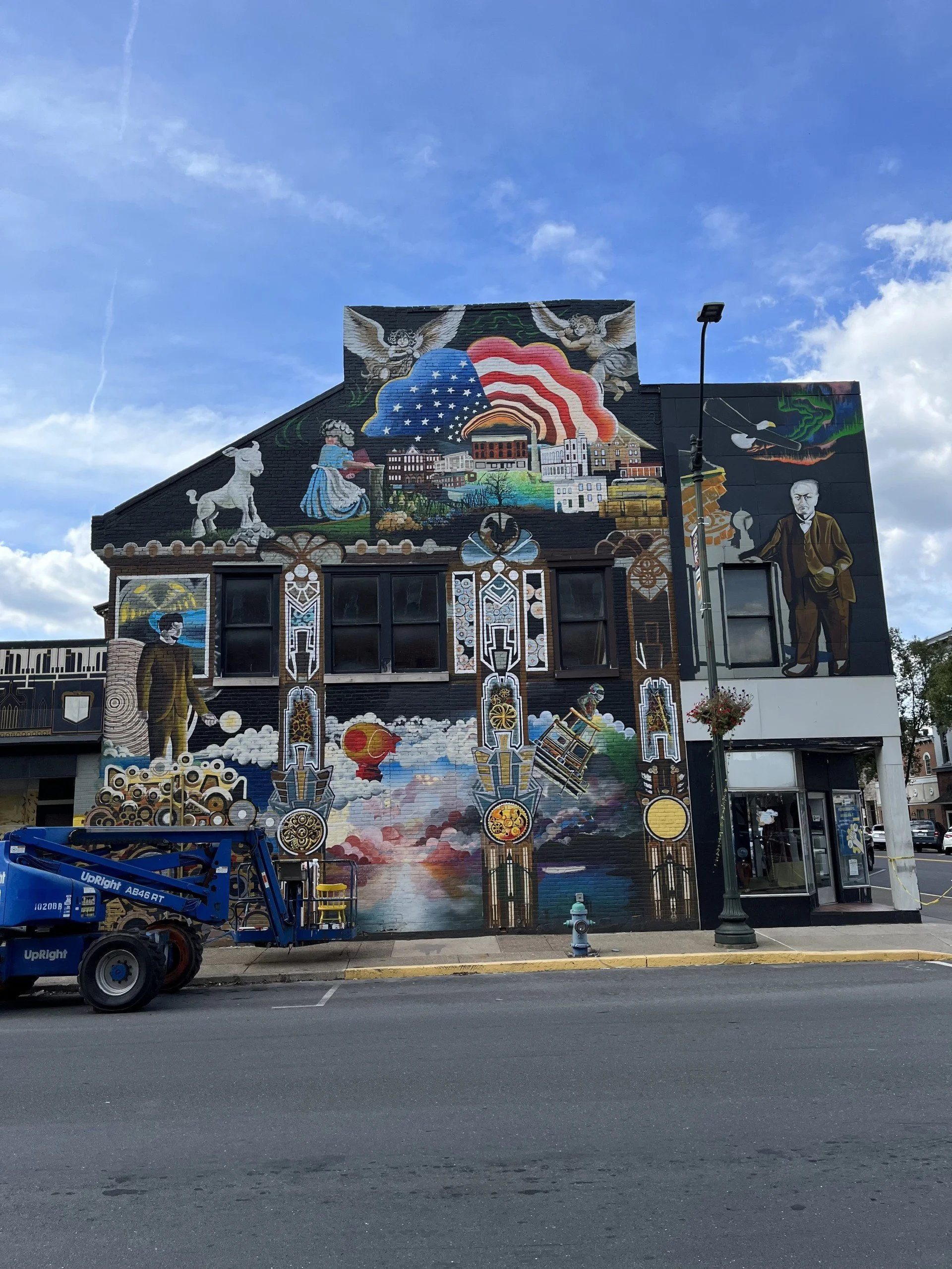 A colorful mural on the upper part of a building depicting symbols of American history and culture, including American flags, a man in period clothing, a Native American figure, and various historic buildings. There are trucks and a street lamp in front of the building under a partly cloudy sky.
