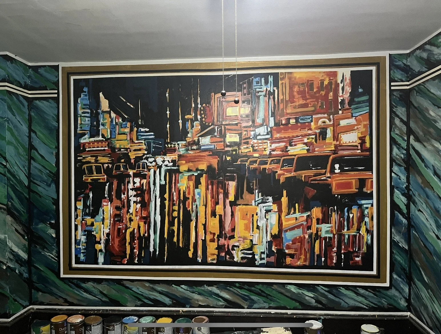 A colorful abstract painting of a city skyline hangs on a wall with a green and black pattern. Paint cans are on the floor below.