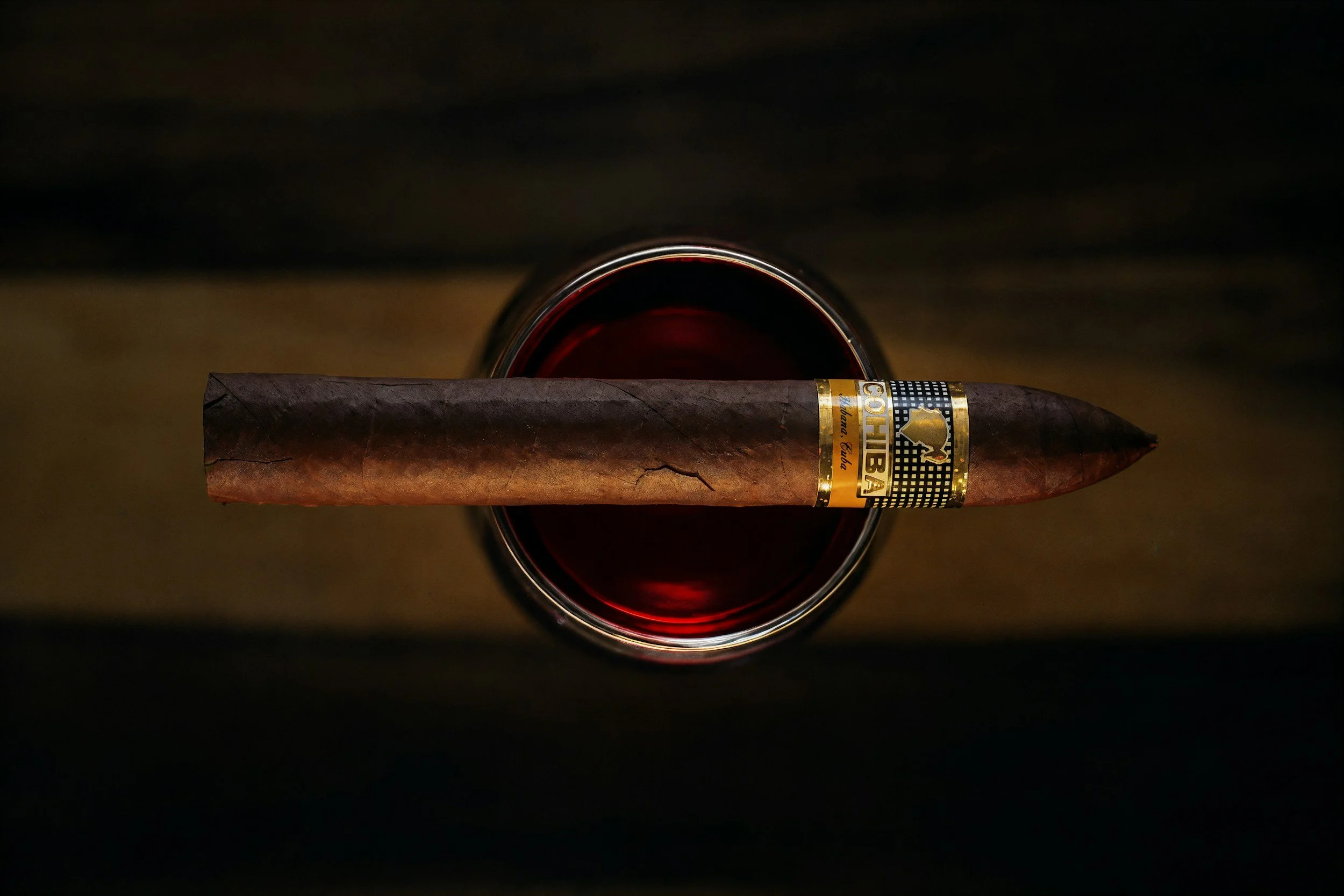 A cigar resting on the rim of a glass of dark red wine.