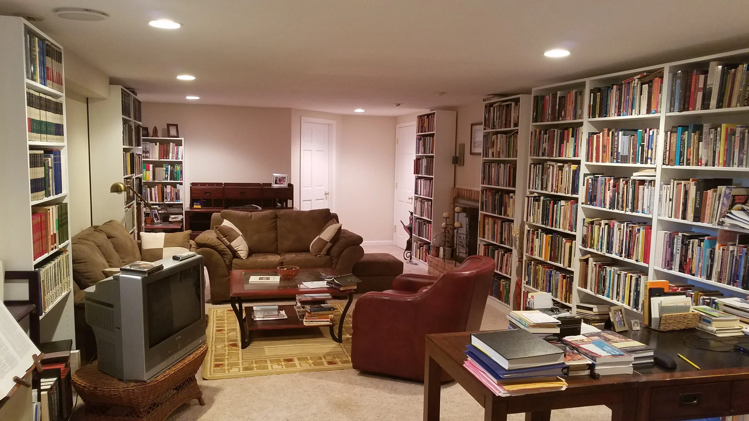 A cozy basement living room filled with tall white bookshelves packed with books on all sides. There are brown and red couches arranged around a wooden coffee table, with a yellow patterned rug underneath. An old television with a remote on top is in the foreground, and a wicker basket is nearby. The back wall has a small desk, framed pictures, and a closed white door. Additional books and small decorative items are on various surfaces, creating a warm, inviting space for reading and relaxing.