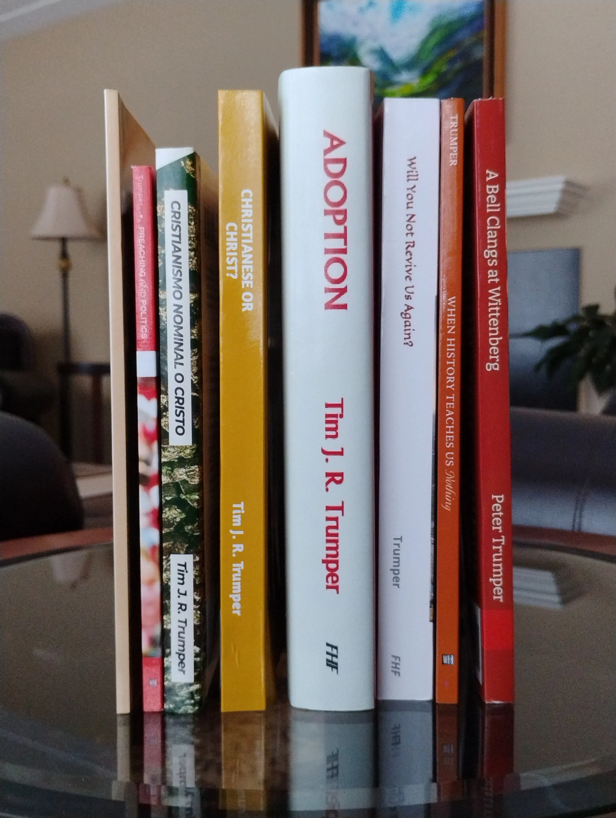 A row of books on a shelf, with titles including 'Aortic Tamponade,' 'Will You Not Revive Us Again?,' and 'A Bell Clangs at Wittenberg,' with a lamp and painting in the background.