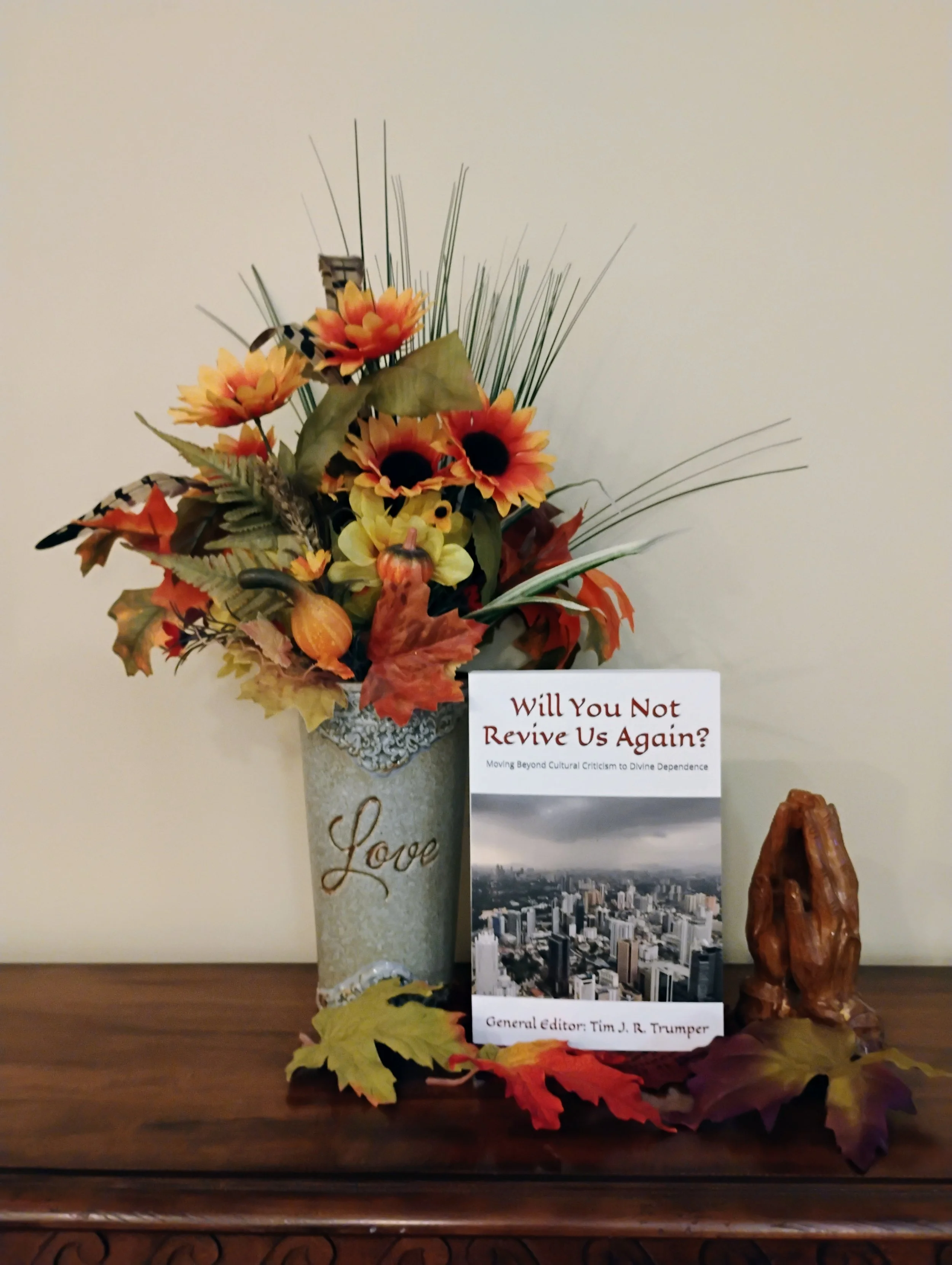 A decorative corner arrangement with a beige vase with 'Love' written on it, containing artificial yellow and orange flowers, leaves, and butterflies, accompanied by a small cityscape photograph and a wooden sculpture, with autumn leaves scattered around.