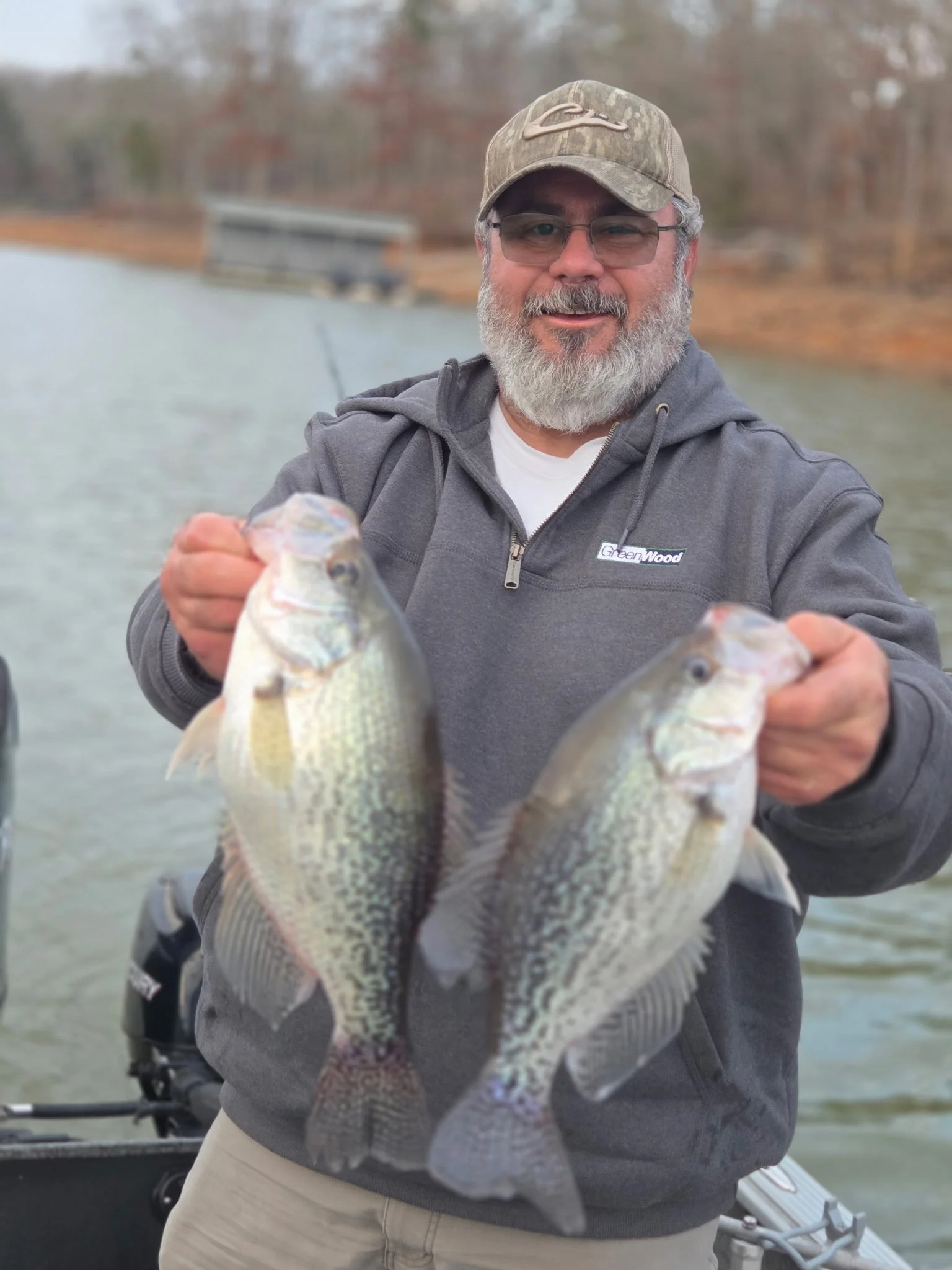 A man on a boat holding up two fish he caught, with a lake and trees in the background.