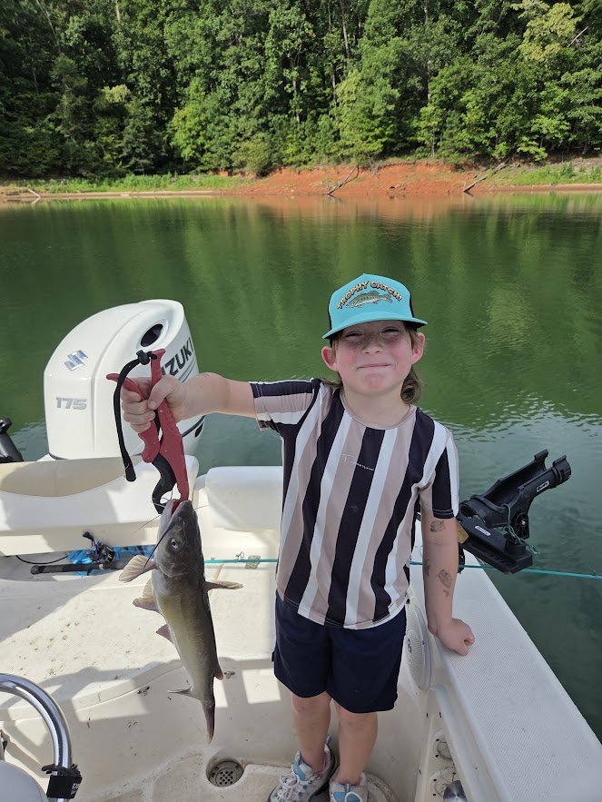 A young boy on a boat holding a fish with a fishing grip, next to a fishing motor, with water and trees in the background.