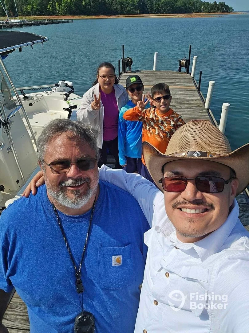 Group of five smiling people taking a selfie on a dock by a lake with a boat and trees in the background.