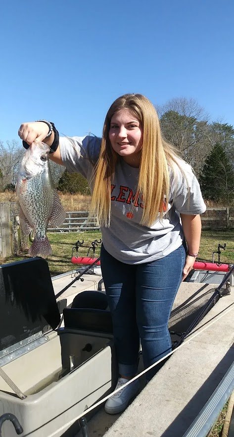 A girl with long blonde hair holding a fish she caught during a fishing trip, standing in a small boat outdoors on a sunny day.