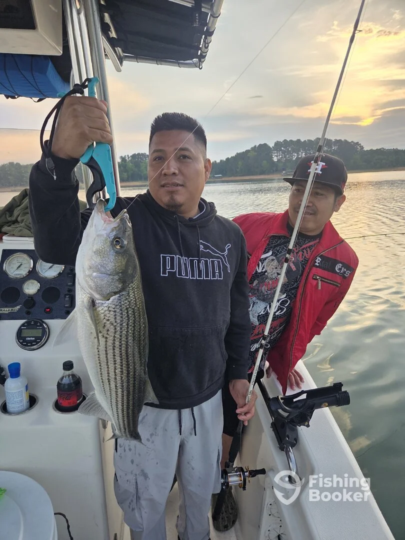 A man holds up a largemouth bass with a fish grip on a boat, with another man in a red jacket and black cap behind him, holding a fishing rod, by a lake at sunset.