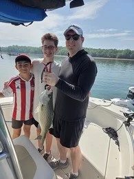Three people on a boat, holding a fish, with a water body and trees in the background. One child wears a red and white striped shirt, and two adults wear black clothing and sunglasses.
