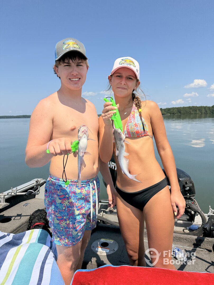 A young man and woman standing on a boat holding fish they caught, with water and a distant shoreline under a clear, sunny sky.