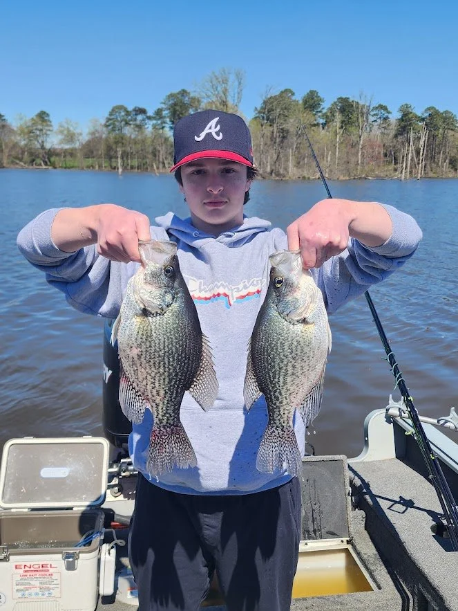 Young boy in a gray hoodie and black cap holding two large fish, standing on a boat by a lake with trees in the background.