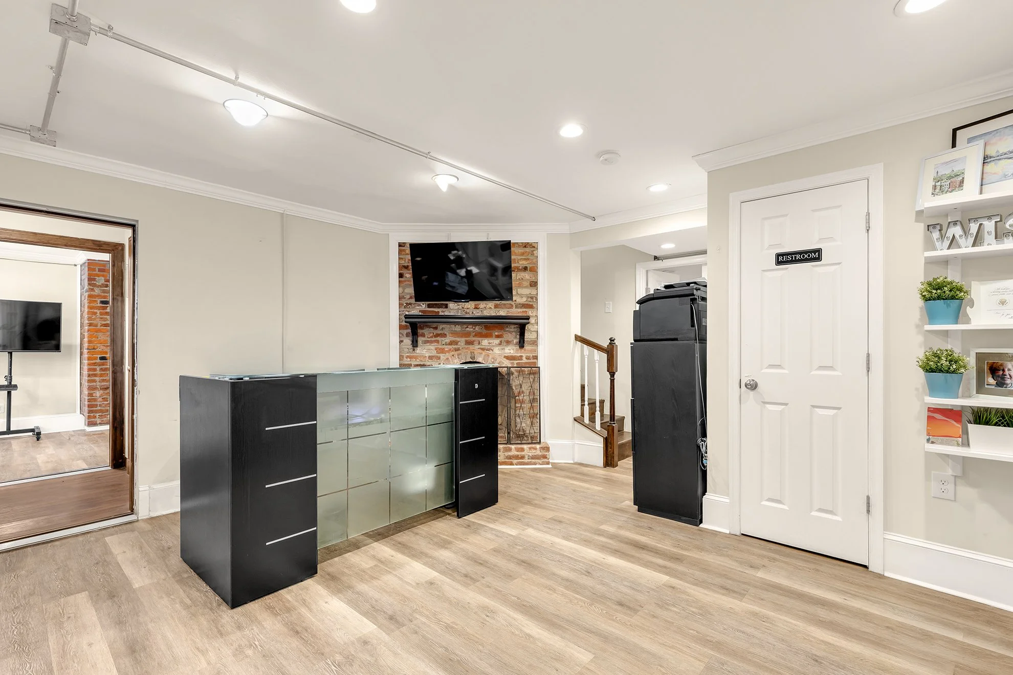 Interior view of a room with a brick fireplace, a mounted flat-screen TV, a black and glass cabinet, a white door labeled "Restroom," and a white shelf with green potted plants and framed pictures. The room has crown molding and wood flooring.