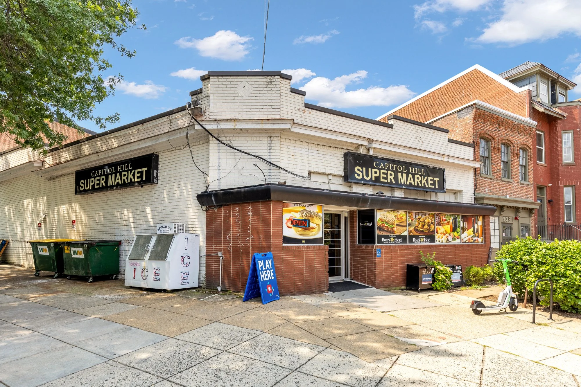 A small grocery store named Capitol Hill Super Market located in a brick and white building on a city street. The storefront has signs with the store's name, a display window showing food images, and a blue sandwich board sign reading 'Play Here.' Th