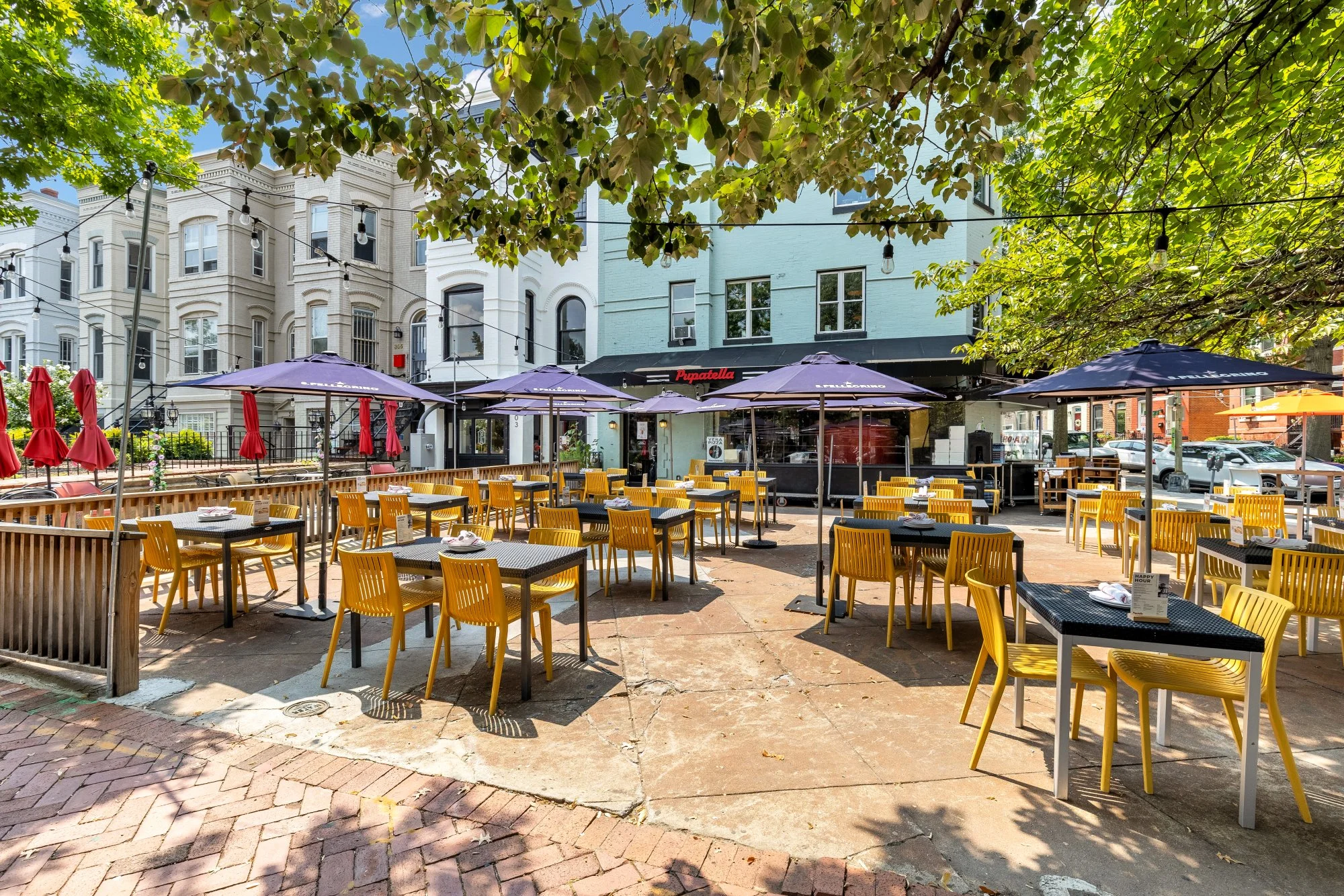 An outdoor restaurant patio with yellow chairs and black tables with white plates, under purple umbrellas on a sunny day, with storefronts and buildings in the background, shaded by trees.