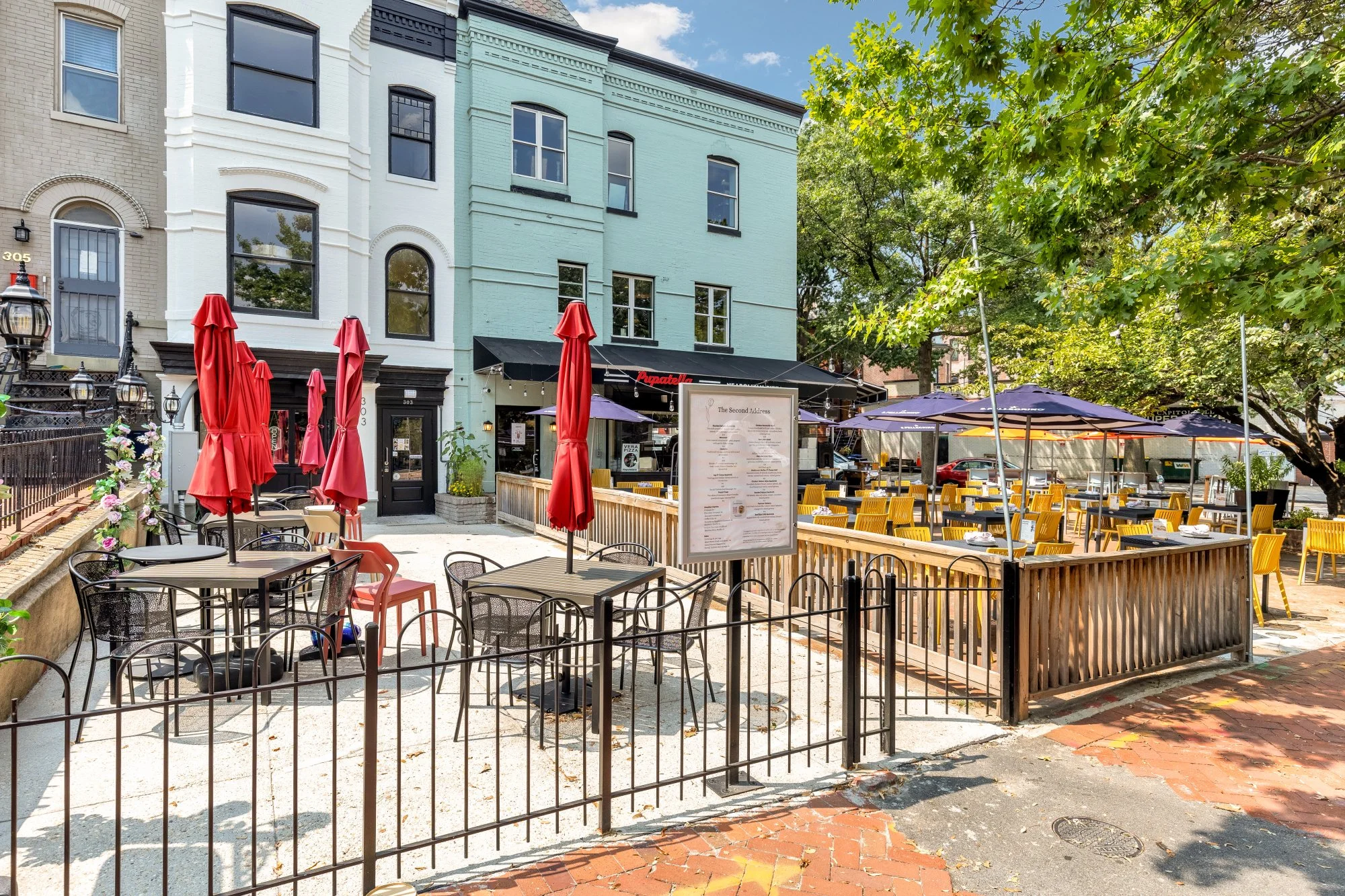 An outdoor restaurant patio with yellow chairs and purple umbrellas, surrounded by a black metal fence, with buildings and green trees in the background.