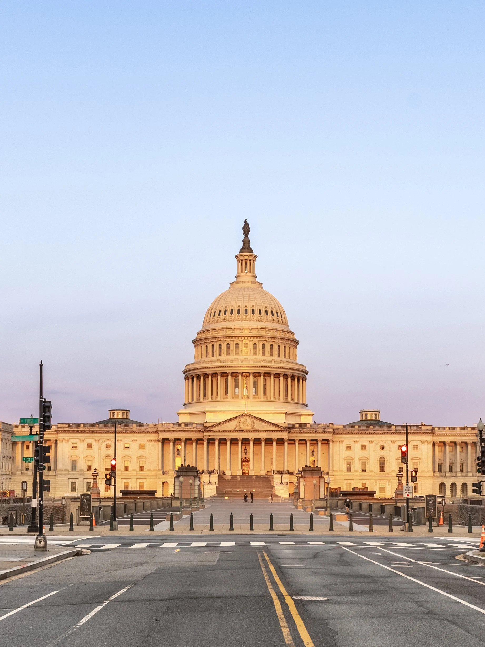 The United States Capitol building at dawn, with a clear sky, traffic lights, and street signs in the foreground.