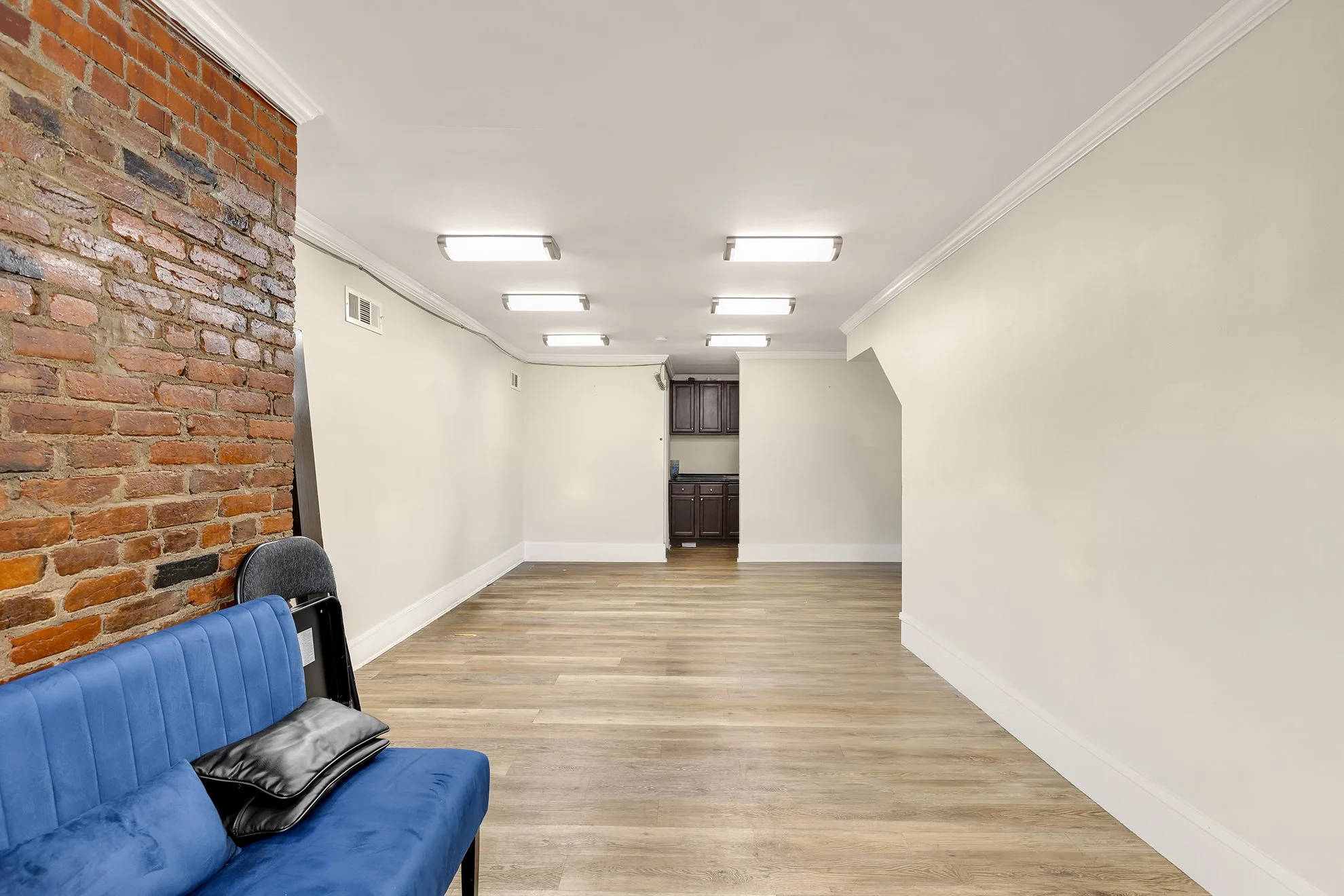 Empty room with white walls, wood flooring, a brick wall on the left, and ceiling lights. There is a blue sofa with black pillows and a small section of additional furniture in the background.