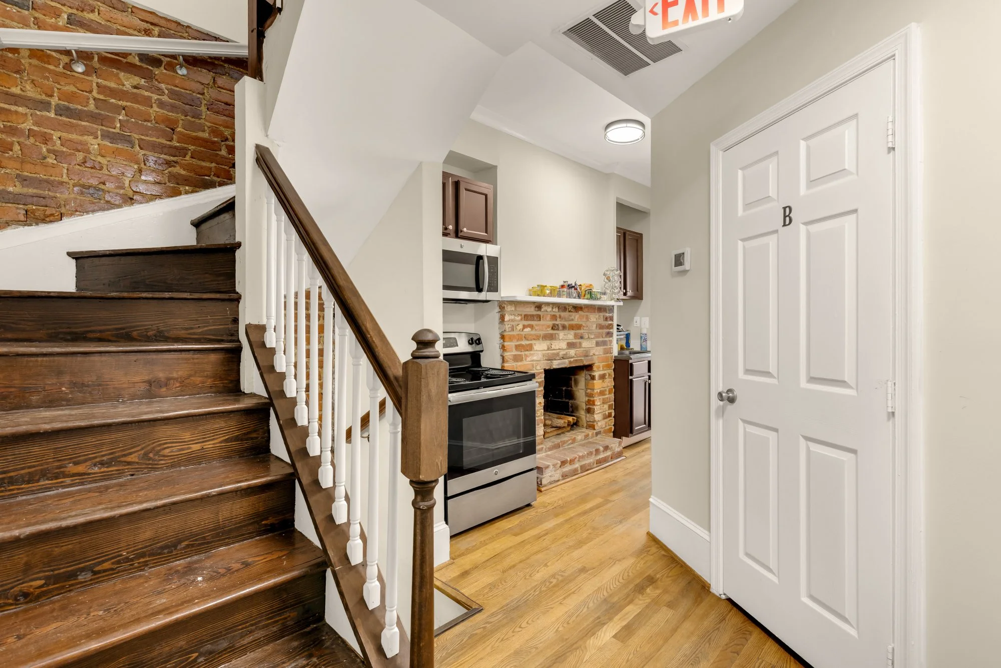 Interior view of a small kitchen area in a residential space, featuring a brick fireplace, a stainless steel stove, wooden cabinets, and a spiral staircase with wooden steps and white balustrade. The ceiling has a round light fixture and an emergency exit sign.