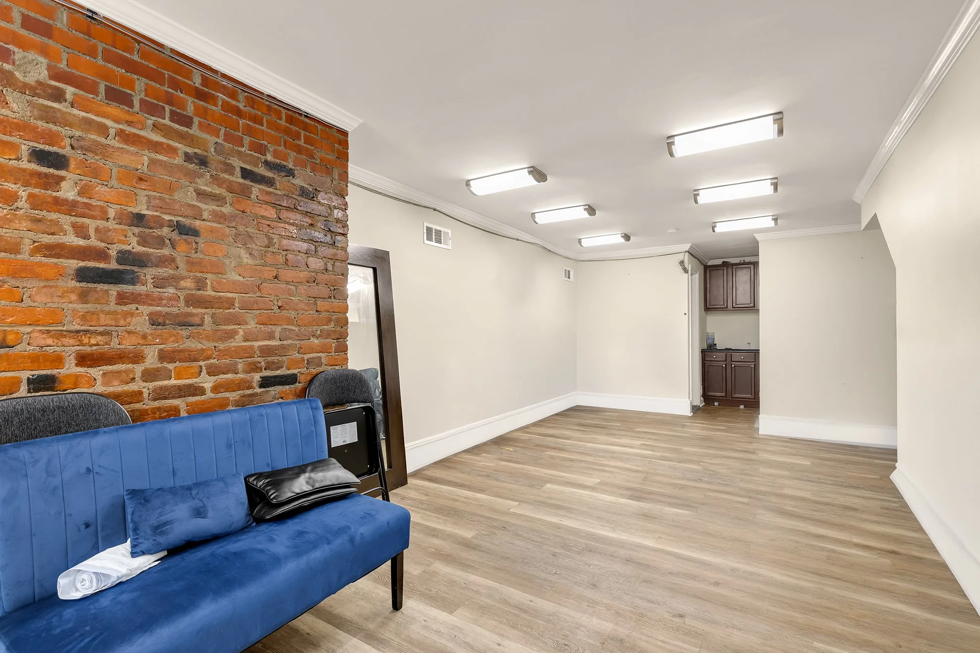Empty room with brick and white walls, ceiling lights, wood floor, a blue couch with a pillow, and some chairs.