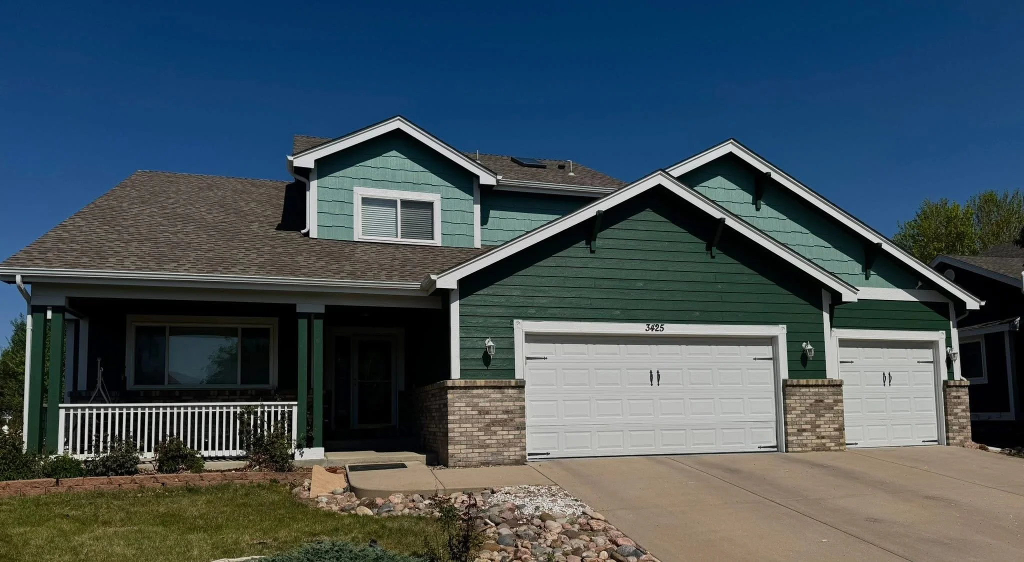 A two-story house with green siding, white garage door, and front porch, under a clear blue sky.