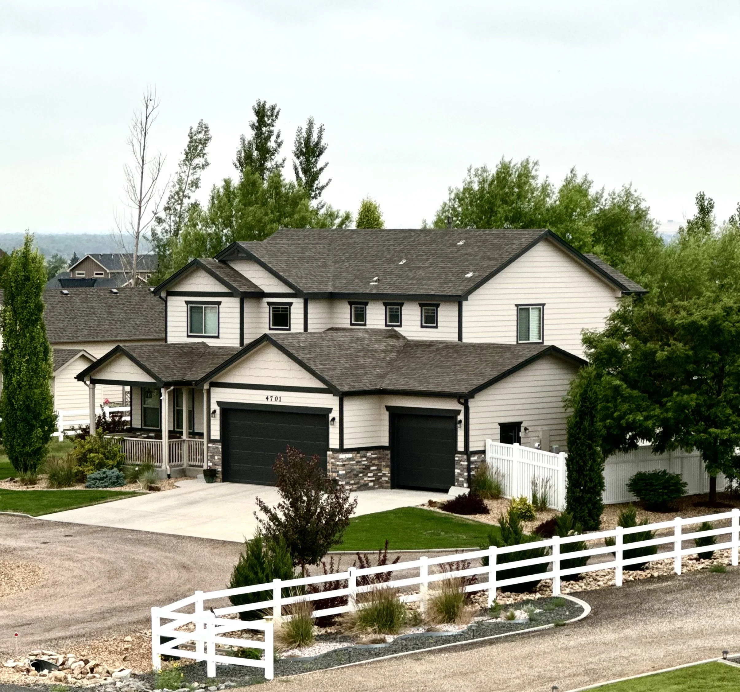 A white modern house with black trim, two black garage doors, surrounded by a white picket fence, with a landscaped yard and trees in the background.