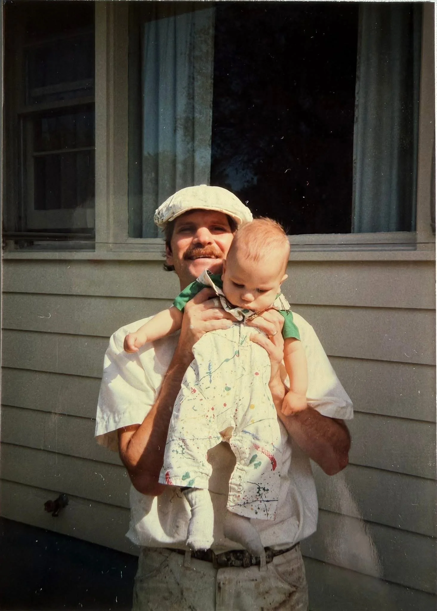 A man with a beard wearing a painter's cap, holding a young child. The man is dressed in a white paint-splattered shirt and khaki pants, standing outside in front of a house with gray siding and windows. The child, wearing a similar white shirt with paint splatters, is looking down.