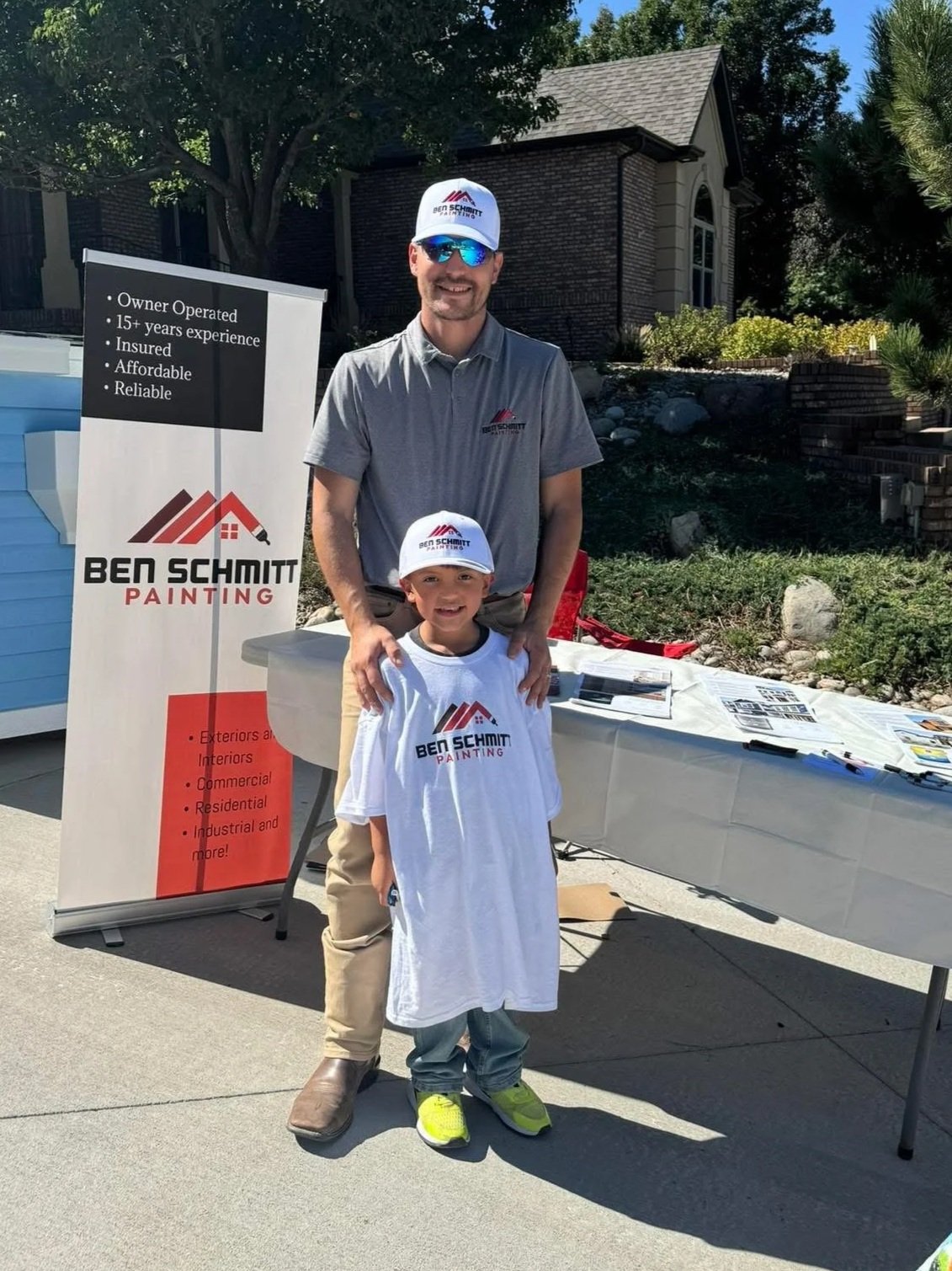 A man and a young boy standing together outdoors in front of a promotional table and banner for Ben Schmitt Painting. They are both wearing hats and shirts with the company's logo. The man is smiling and has his arm around the boy. The banner lists services offered by the company, including exterior, interior, commercial, residential, and industrial painting.
