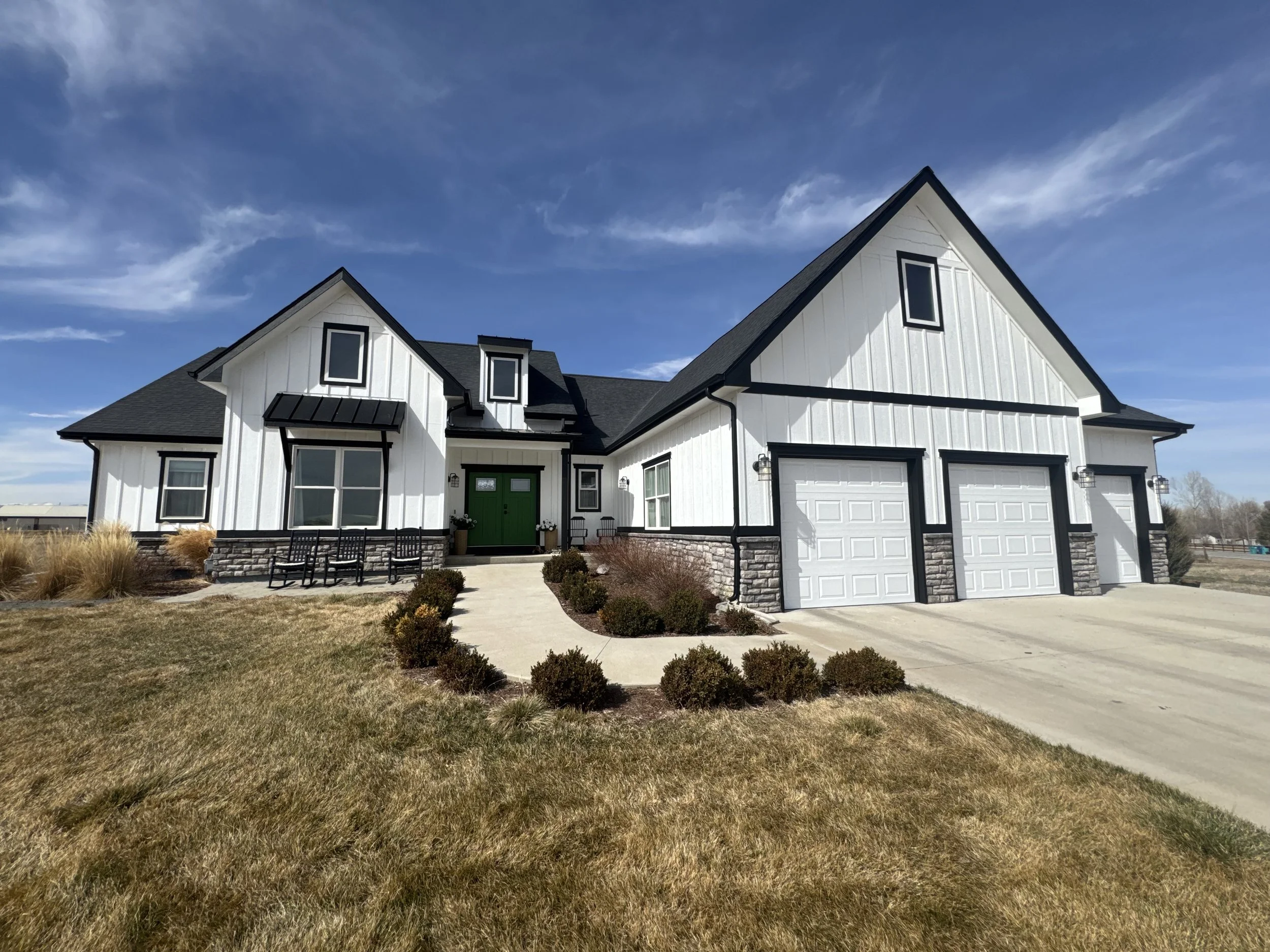 Modern two-story house with white exterior walls, black window frames, and a green front door. It has a two-car garage and a front porch with black benches. The yard has dry grass with some bushes, a concrete pathway, and outdoor wall lights. The sky is partly cloudy with a blue background.