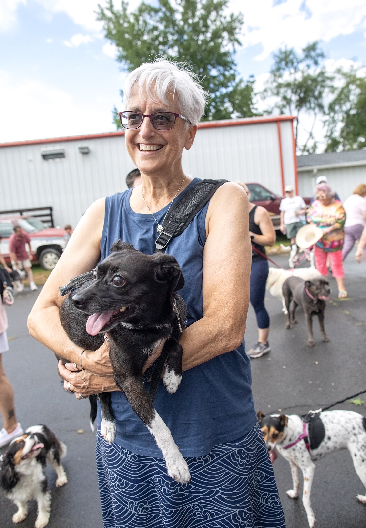 A smiling older woman with short white hair and glasses holding a black and white puppy at an outdoor event with other dogs and people in the background.