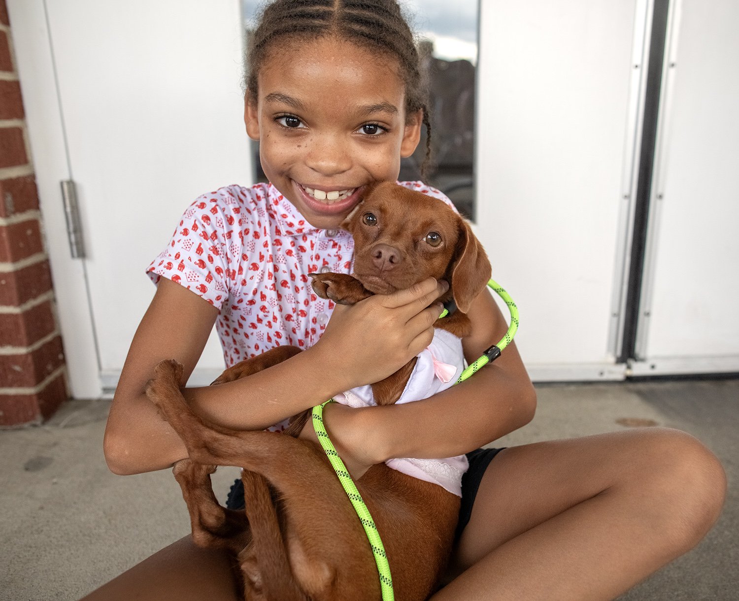 A young girl with a big smile is holding a brown puppy. The girl has braided hair and is wearing a white shirt with red heart patterns. She appears happy as she hugs the puppy, which has a green leash and is wearing a white harness with a pink bow. They are sitting outside, near a white door with a brick wall on the side.